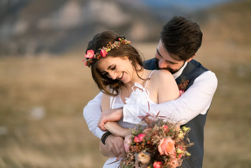 Braut mit Blumenkranz lacht, der Bräutigam umarmt sie von hinten, auf einer herbstlichen Bergwiese