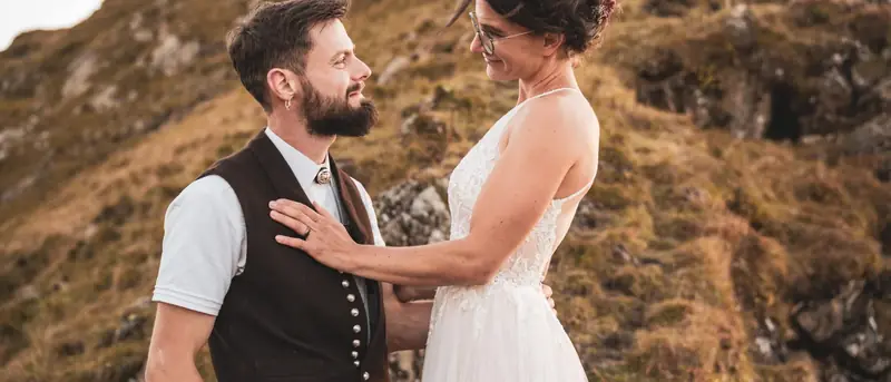 Braut und Bräutigam in Tracht stehen sich an einem sonnigen Berghang gegenüber, Bild im Kinoformat