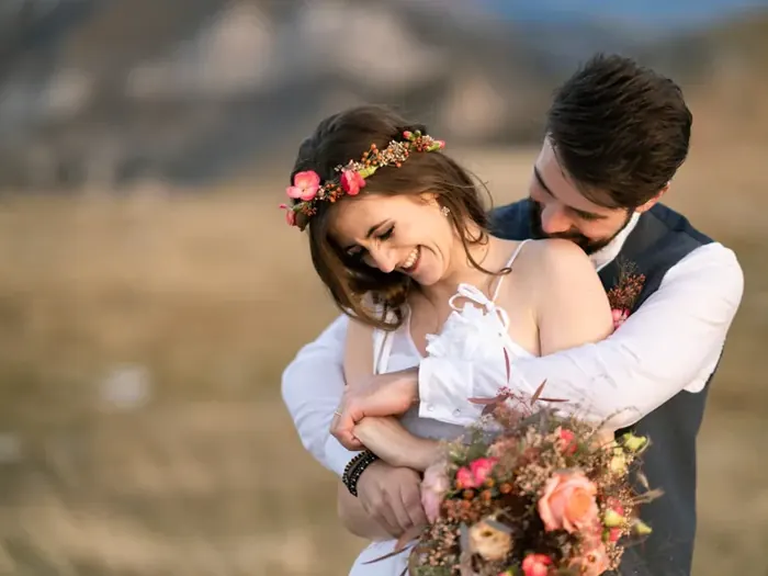 Braut mit Blumenkranz lacht, der Bräutigam umarmt sie von hinten, auf einer herbstlichen Bergwiese