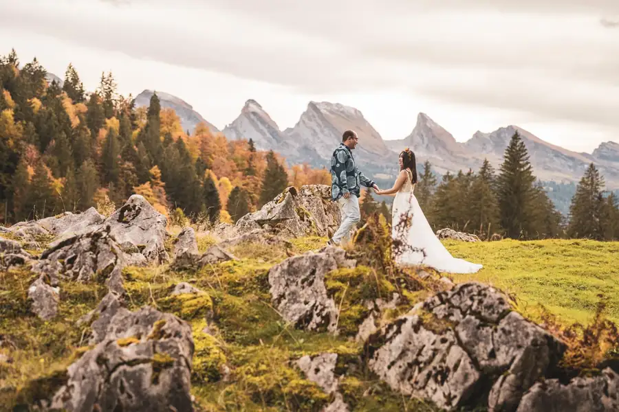 Brautpaar geht Hand in Hand über eine Alpweide mit Felsen, dahinter die Churfirsten im Herbst